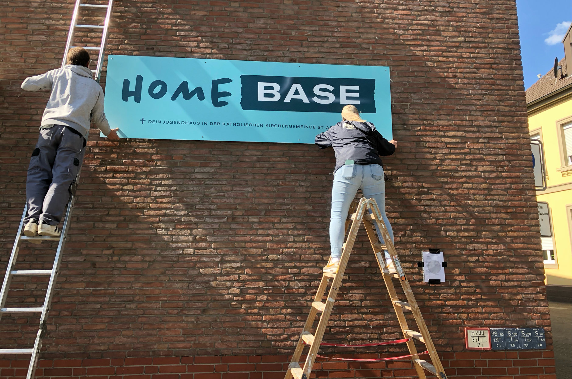 Zwei Handwerker montieren das neue Schild des Jugendhauses Homebase an die Fassade